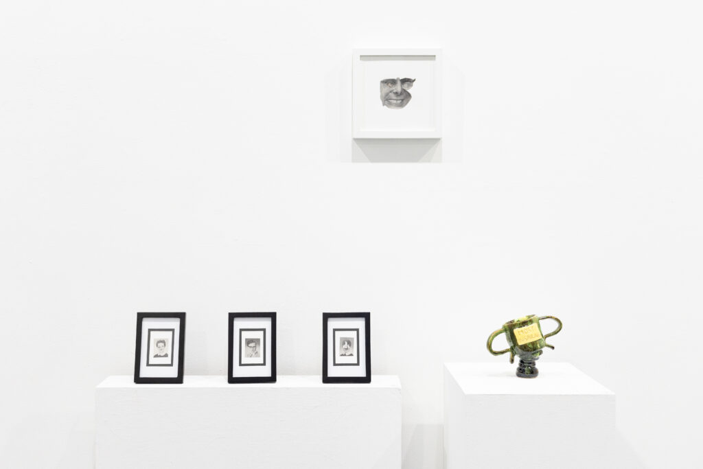 Three small framed portrait drawings sit on a white plinth beneath a larger framed face drawing, with a green ceramic trophy displayed on a separate pedestal