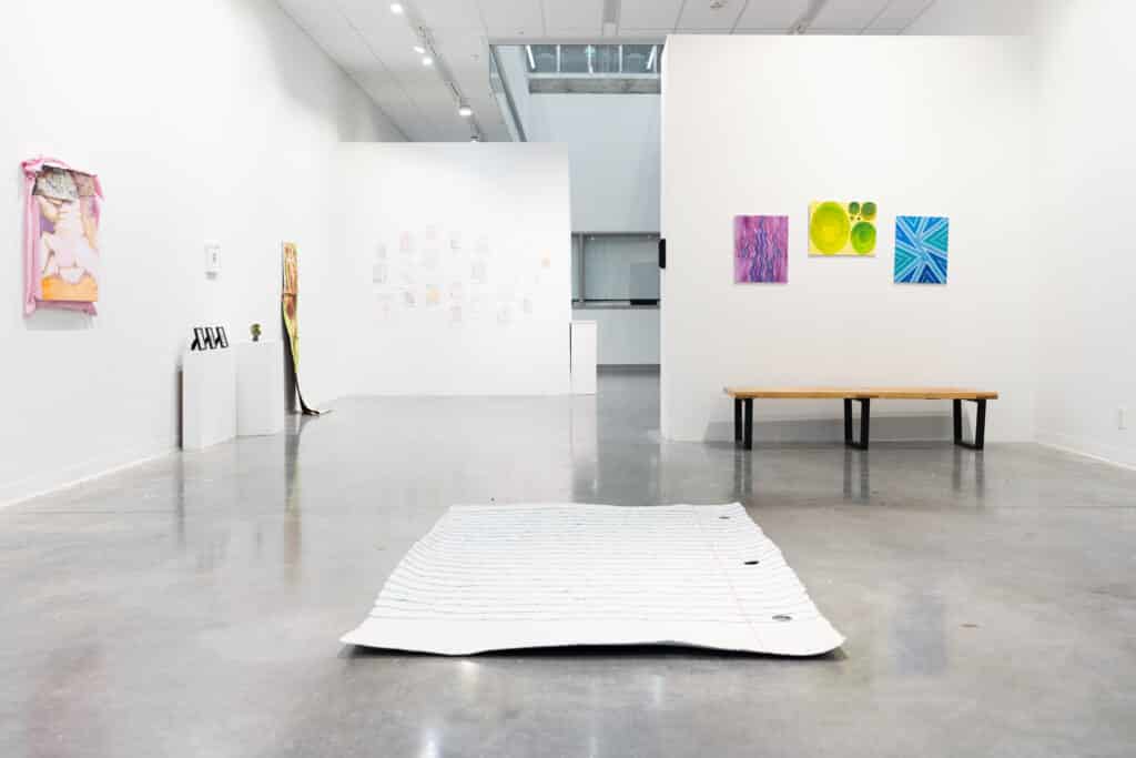 A bright gallery space shows multiple small artworks on white walls, a wooden bench to the right, and a large lined paper sculpture laid flat on the floor in the foreground.