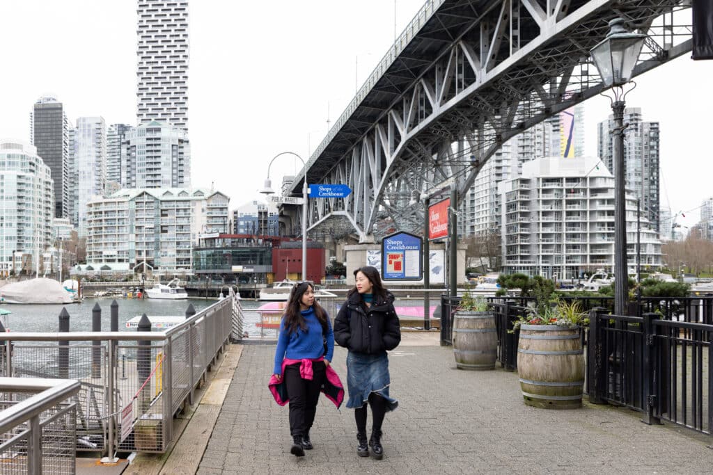 Two people stroll along a waterfront promenade beneath a large bridge, mid-conversation, with boats, buildings, and soft grey skies stretching around them.