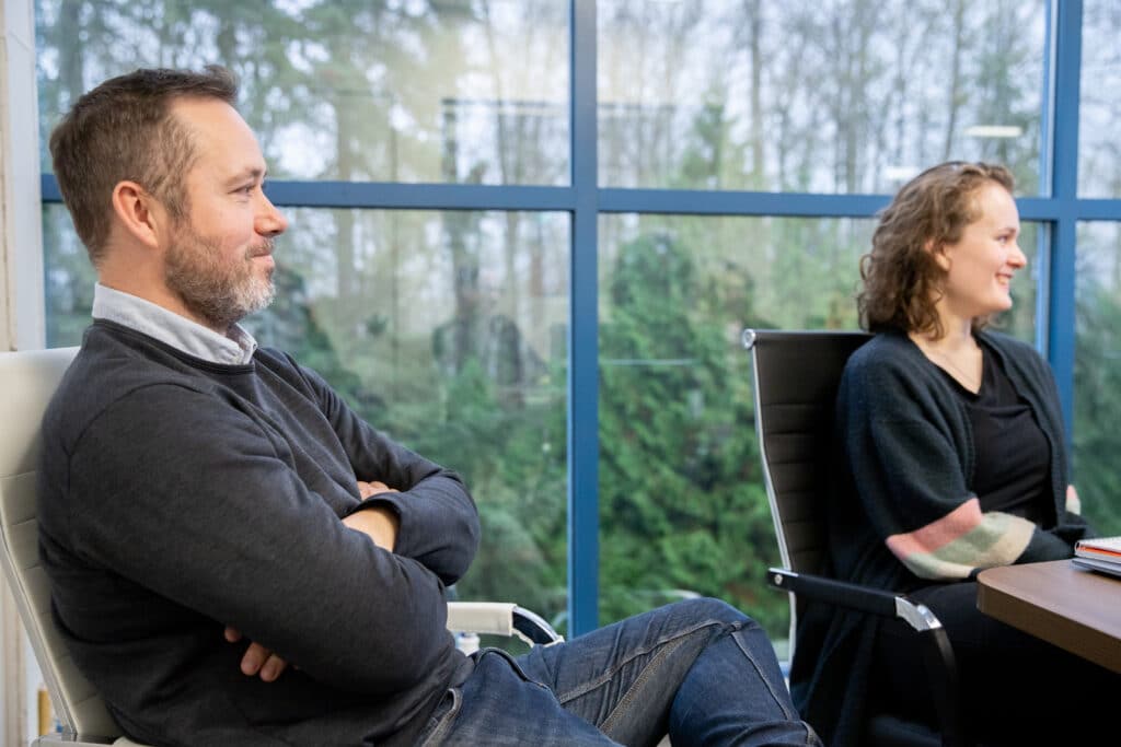 Two people sit in a meeting room with large windows, listening attentively during a discussion.