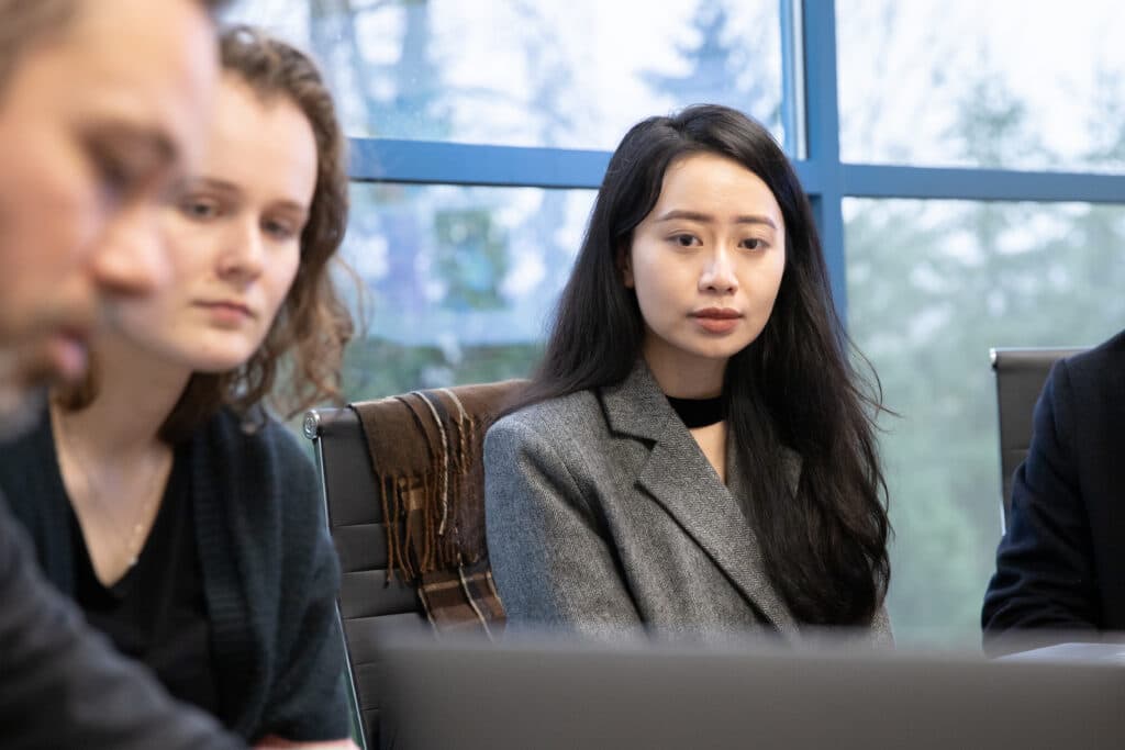 Three people sit at a meeting table, focused on a laptop during a group discussion.