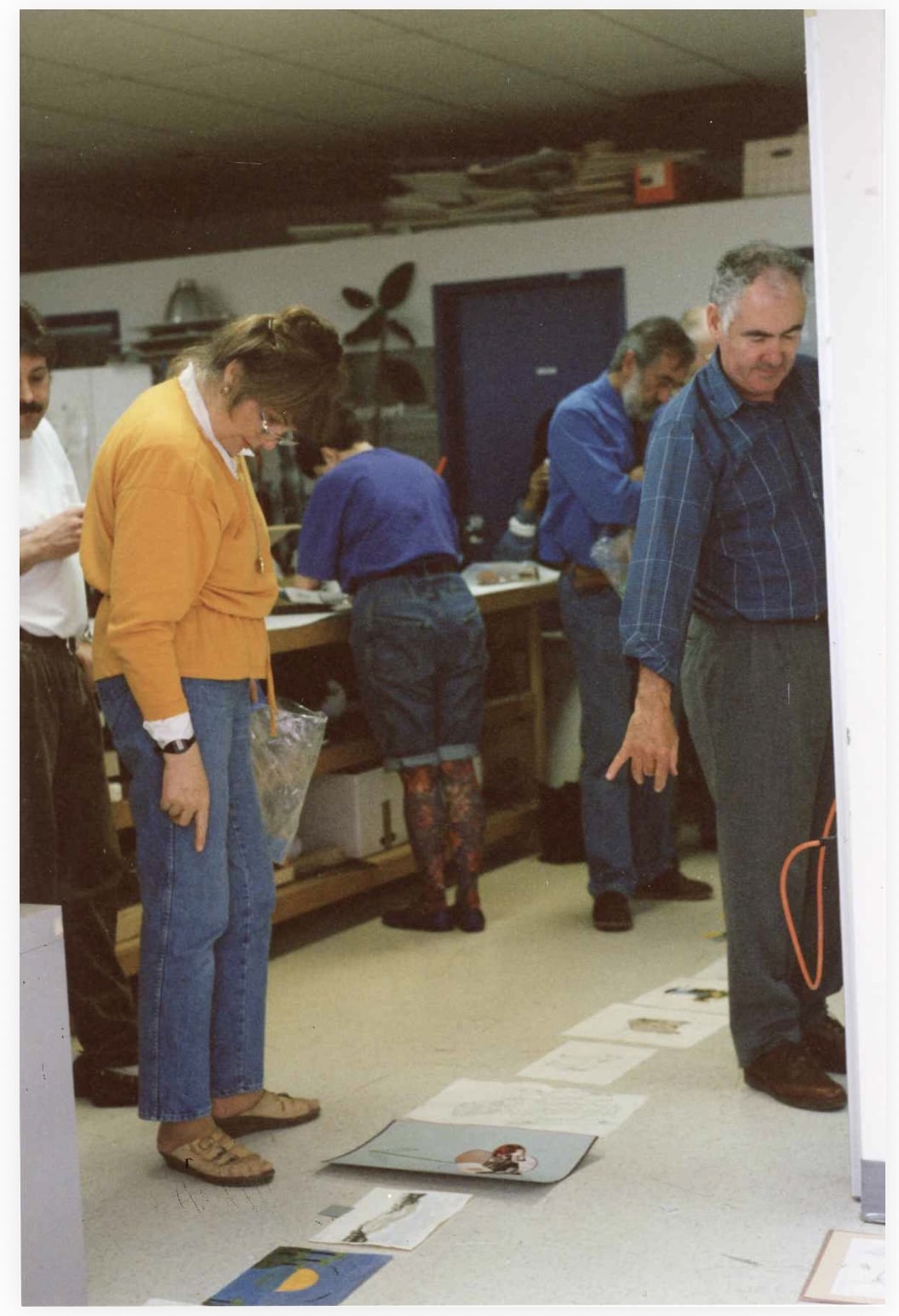 People stand in a studio space looking down at artworks laid out on the floor while others work at tables in the background.