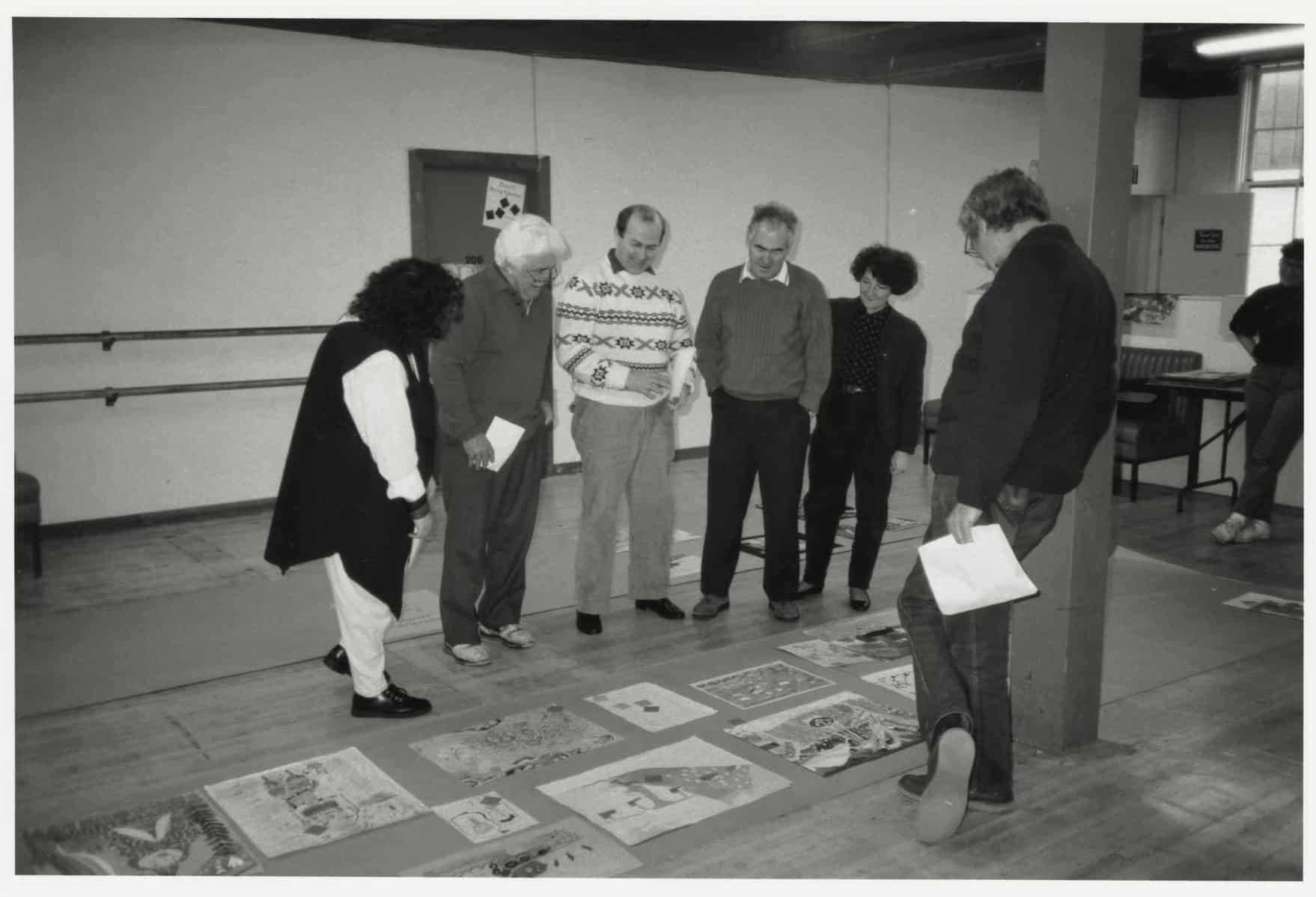 Group of people stand in a room looking down at children’s artworks arranged on the floor during a review or selection process.