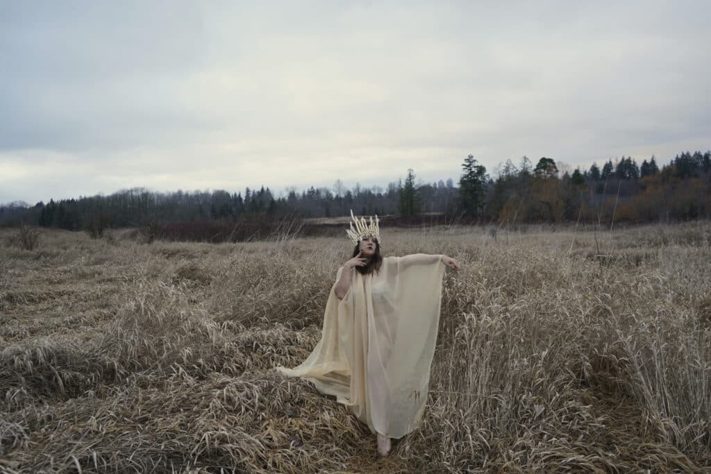 A person moves through a field of pale, dried grass beneath a wide, cloudy sky, wearing a flowing, translucent garment and a sculptural headpiece. Their arms lift gently, as if in dialogue with the land.