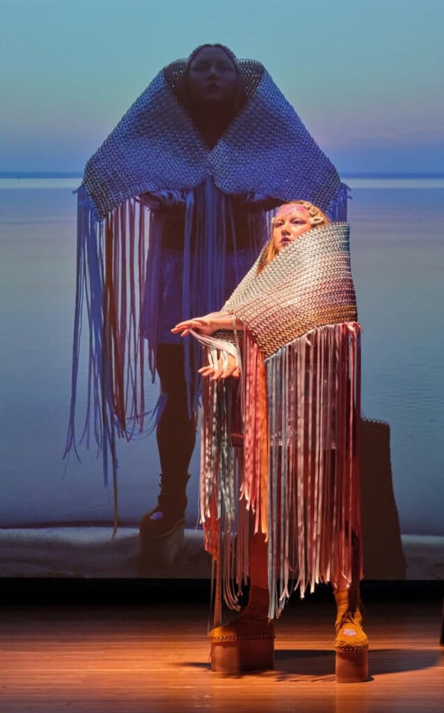A person stands on a stage before a projected shoreline, wrapped in a woven, fringe-lined garment that drapes like a mantle. Their hands extend gently forward, creating a layered image where body and landscape echo one another.