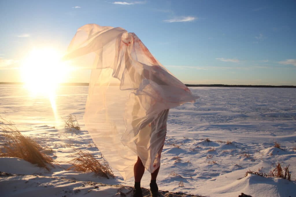 A person stands on a snowy shoreline at sunset, wrapped in sheer fabric that catches the wind and light. The glowing horizon and drifting cloth create a soft, fleeting silhouette against the open sky.