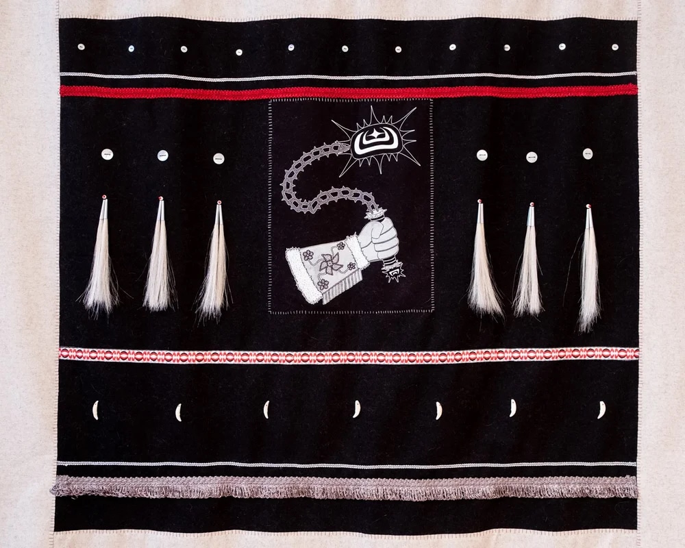 A dark textile panel bordered with red and white stitching features rows of small circular forms and hanging tassels. At the centre, a stylized hand holds a chain-like form beneath a radiant symbol, creating a striking, symmetrical composition.