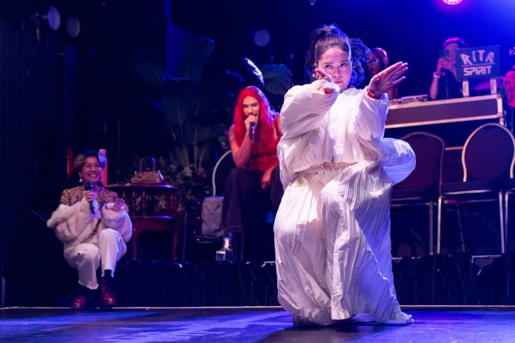 A performer in flowing white kneels at centre stage, arms extended in a sharp, focused gesture, while seated hosts and a DJ booth glow behind them in vivid stage lighting.