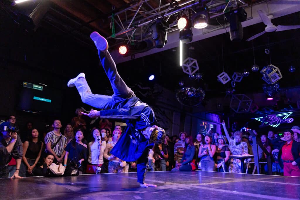 A breakdancer balances on one hand in a powerful freeze, legs extended overhead, as a packed audience surrounds the stage, faces lit by colourful club lights and reflected disco balls.