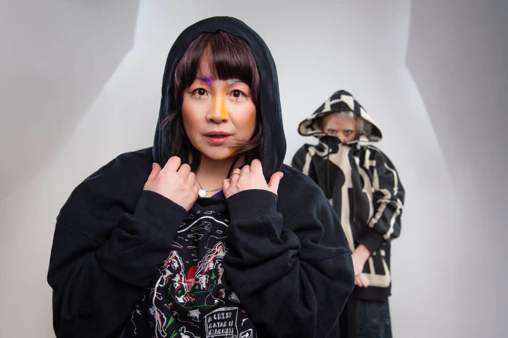 A person in a black hoodie looks directly toward the camera, hands holding the hood near their face, with bold yellow and purple makeup across their nose and brow. Another hooded figure stands behind them in soft focus, creating a layered, slightly mysterious portrait against a clean white background.