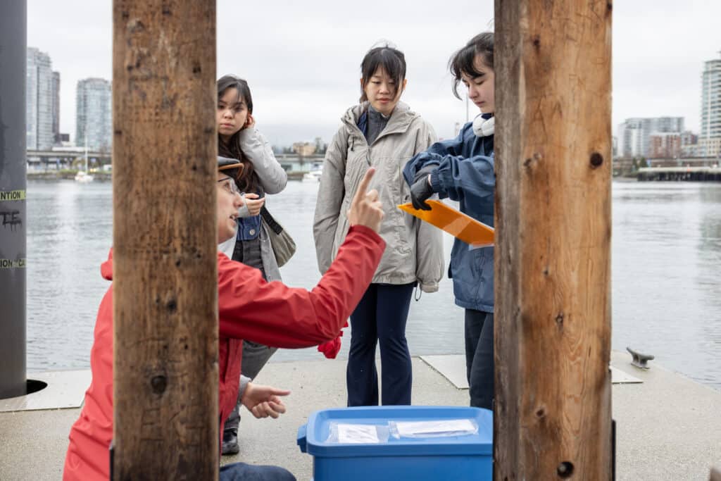 A small group gathers at the water’s edge as one person gestures while explaining, others listening closely; wooden posts frame the scene like a pause in shared learning.