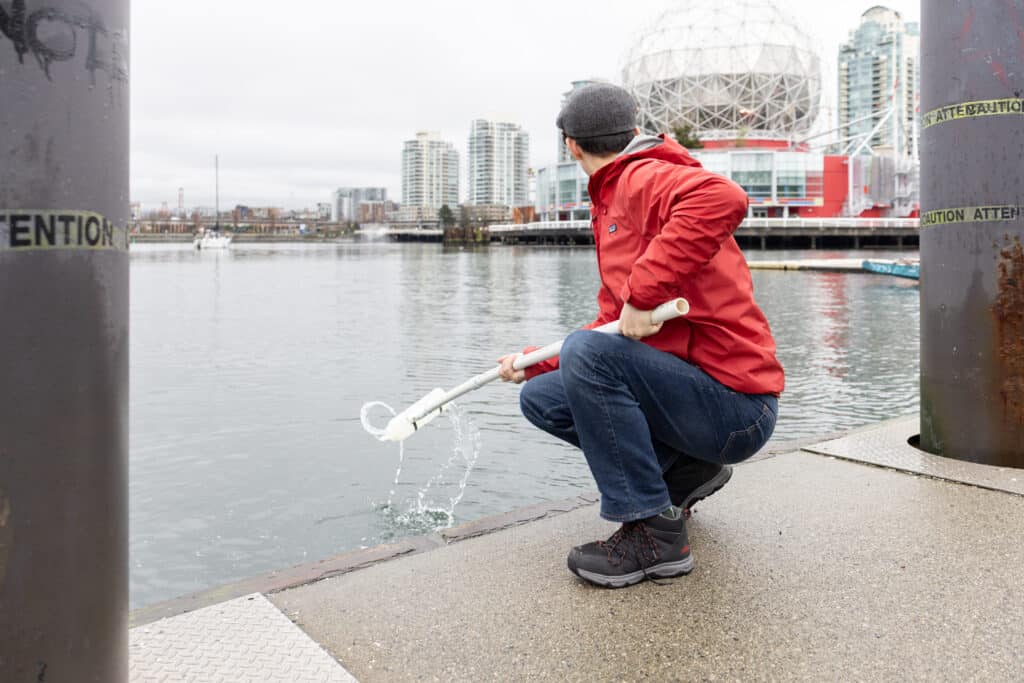 A person kneels by the water, dipping a long tool into the surface, creating a brief splash as the city skyline watches from across the inlet.