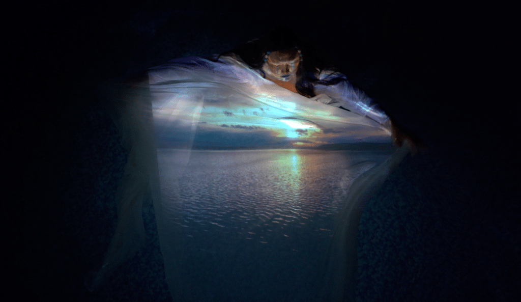 A person emerges within a deep blue scene, their flowing garment reflecting a horizon of water and light. The image blends body and landscape, as if the figure is carrying the sunset within them.