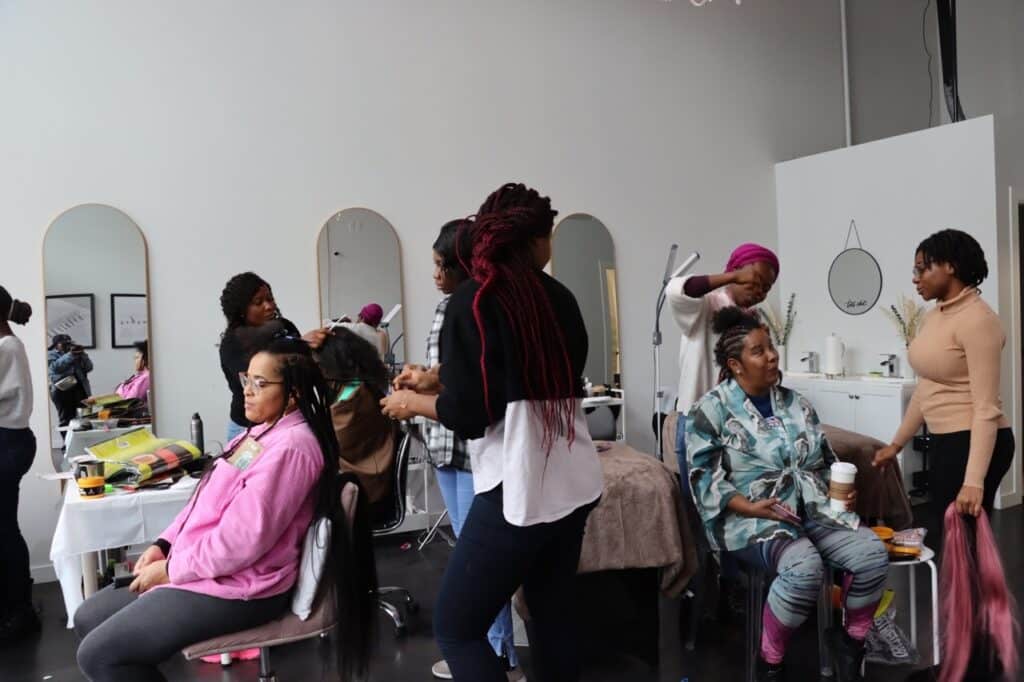 In a bright studio filled with mirrors and soft light, several people gather around styling chairs, braiding and arranging hair while others sit and talk. The room hums with quiet concentration, care, and shared conversation.