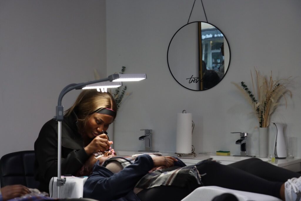 In a bright studio filled with mirrors and soft light, several people gather around styling chairs, braiding and arranging hair while others sit and talk. The room hums with quiet concentration, care, and shared conversation.