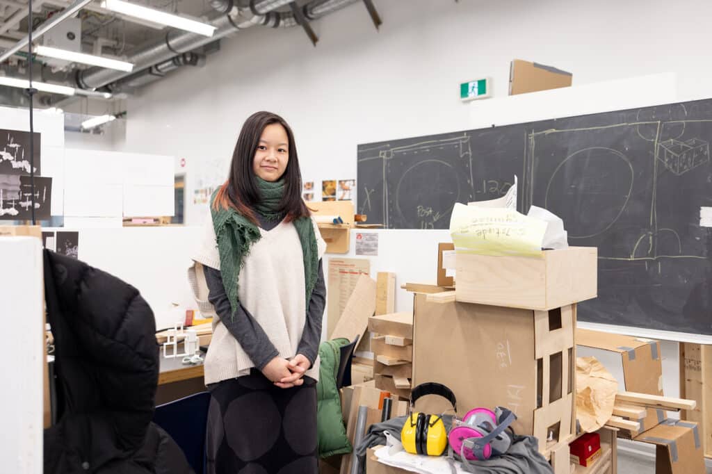 A person stands in a bright design studio beside wooden models and sketches on a chalkboard. Surrounded by tools, materials, and worktables, they pause calmly within a space shaped by making and ideas.
