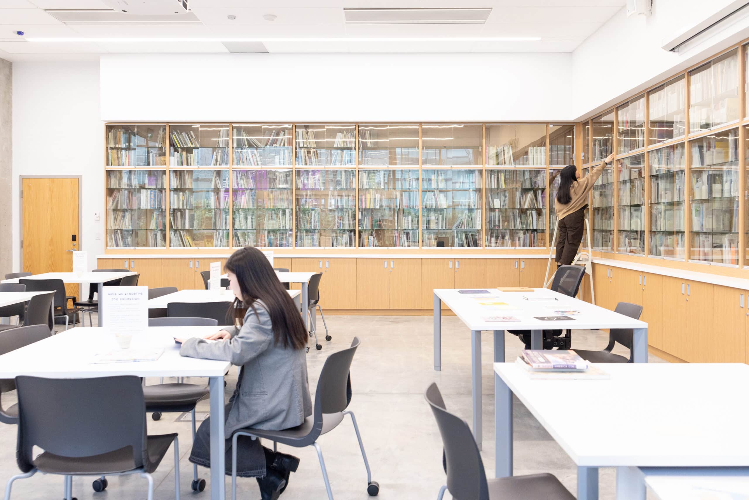 A wall of books behind glass with several tables in front. One person sits at a table looking at a book while another is on a ladder reaching for a book on a high shelf.