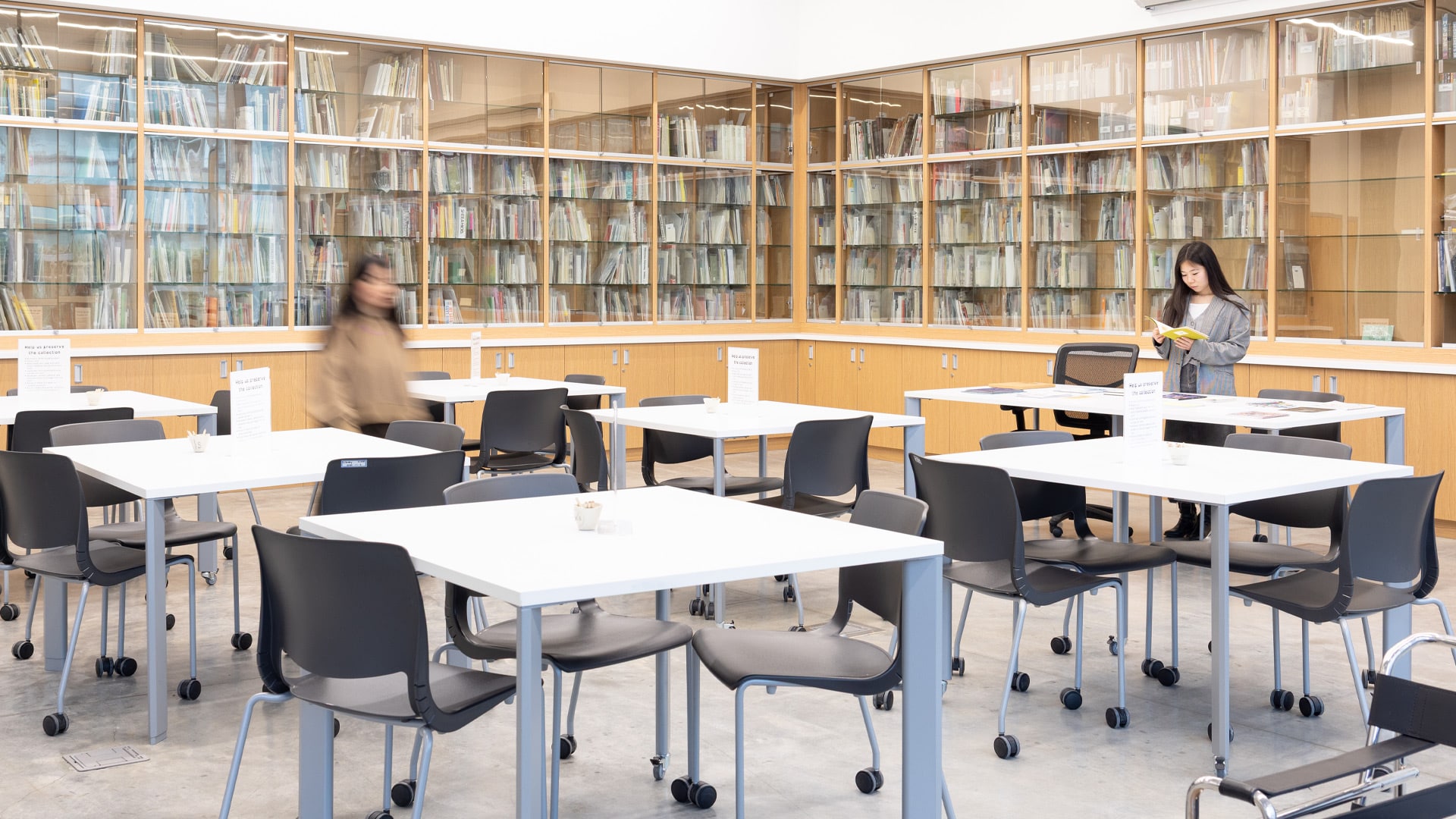 A bright, modern library archives reading room at Emily Carr University with floor-to-ceiling glass-fronted wooden bookshelves filled with books and archival materials lining the walls. White rectangular tables with grey rolling chairs are arranged throughout the space. A student stands reading a book on the right side, while another person walks through the left side of the room. The contemporary space features clean lines and natural lighting.