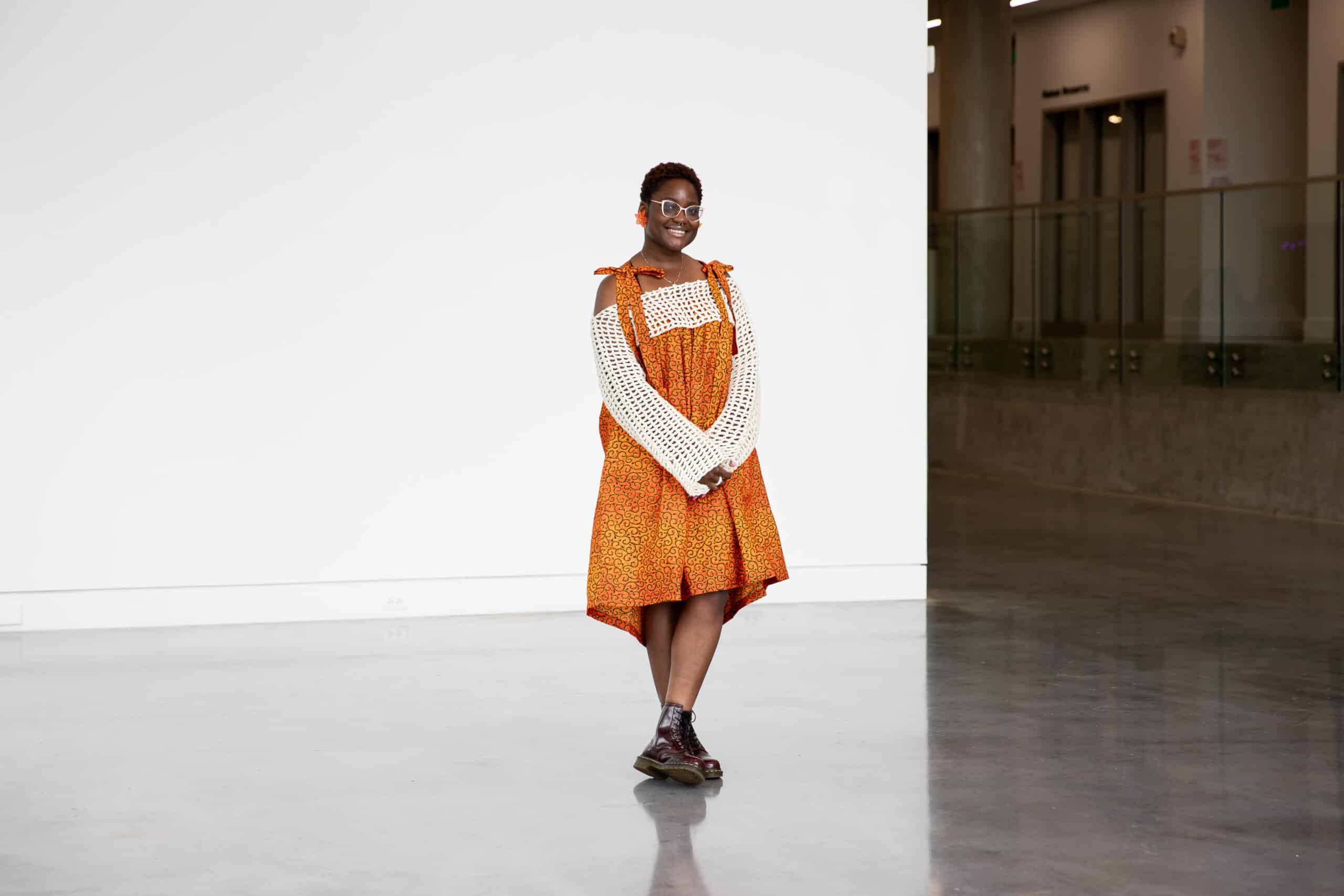 A person stands smiling in a spacious, light-filled interior, wearing an orange patterned dress layered over a cream knit top. Polished floors and a white wall create a calm backdrop as they pause mid-step, hands gently folded.
