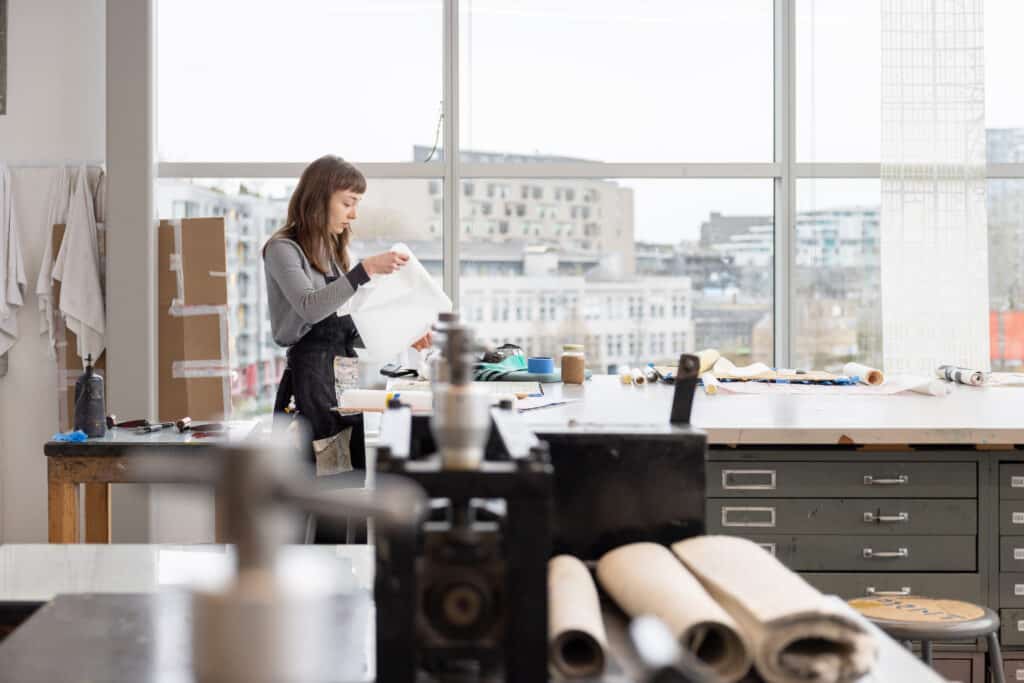 A person stands at a large worktable in a bright studio, carefully lifting a sheet of paper. Tools, rollers, and jars rest nearby, while a wide window opens onto a soft city view, holding a quiet moment of making and focus.