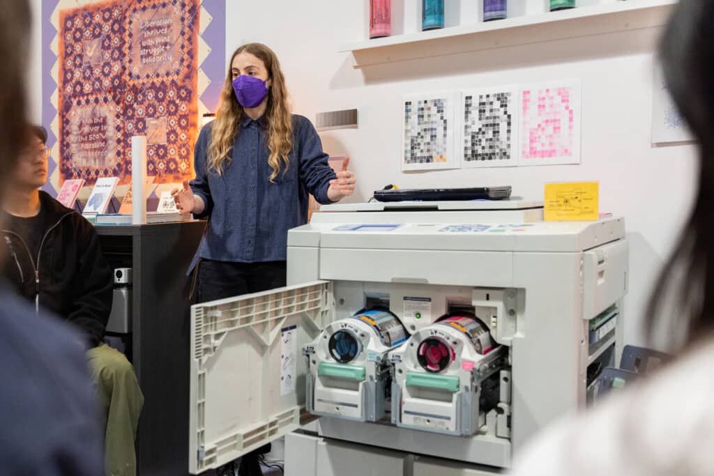 A person stands beside an open printer, explaining a process to a small group. Ink cartridges glow with colour inside the machine, while prints and patterns line the walls behind.