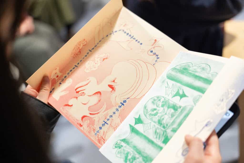 Hands hold open a small artist’s book, its pages washed in soft red and green ink. Dreamlike figures and lines drift across the paper, inviting a quiet, tactile moment of looking and reading.