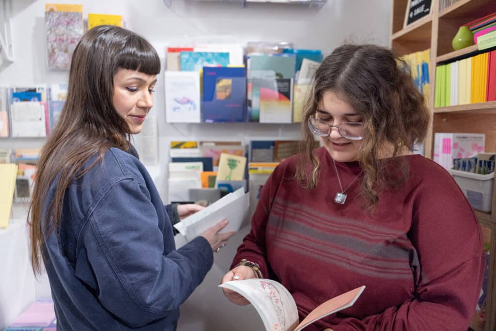 Two people stand close together among shelves of small books, quietly flipping through pages. Their attention rests on the printed details, sharing a calm moment of discovery.