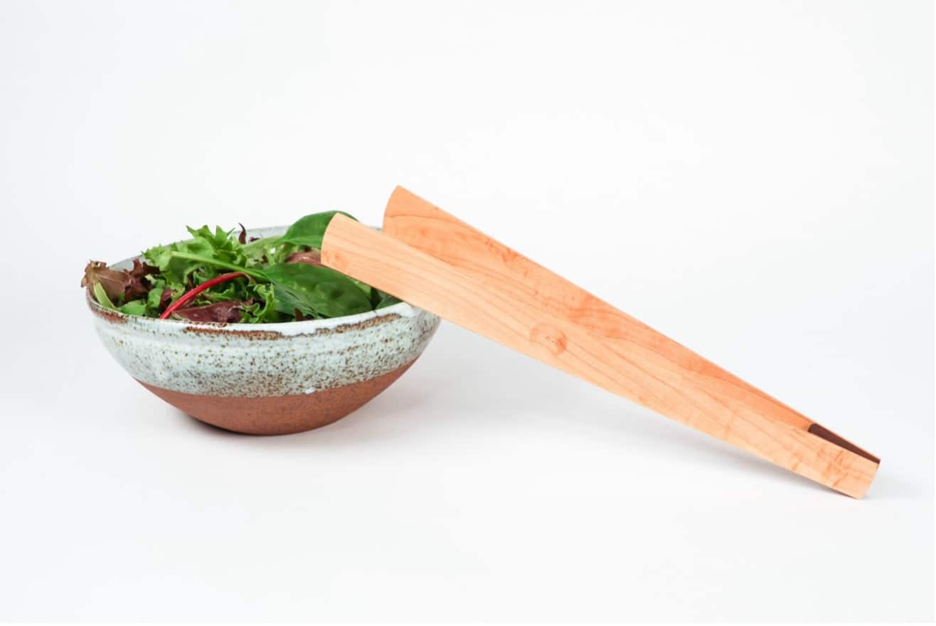 A ceramic bowl filled with fresh leafy greens sits on a clean white surface, beside a long wooden serving utensil with a smooth, tapered shape. The pale wood and soft greens create a simple, quiet moment of preparation.