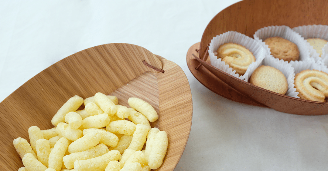 A shallow bamboo dish holds pale, puffed snacks, while a nearby wooden bowl contains neatly arranged cookies in paper cups. The simple wooden forms frame the treats with a soft, natural warmth.