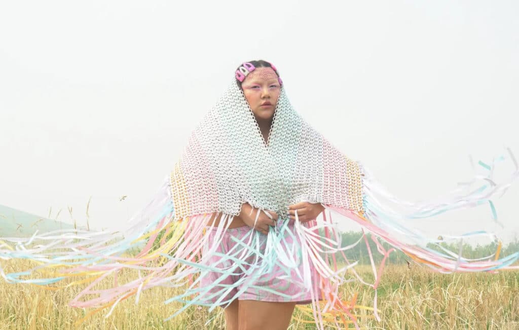 A person stands in a grassy field wrapped in a textured, net-like garment with long, pastel ribbons that trail in the air. The soft sky and open landscape frame a still, grounded presence.