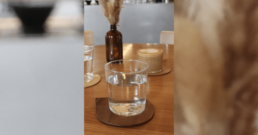 On a wooden table, a clear glass of sparkling water rests on a round wooden coaster. Behind it, a latte and a small amber bottle with dried grasses create a calm café moment in warm light.