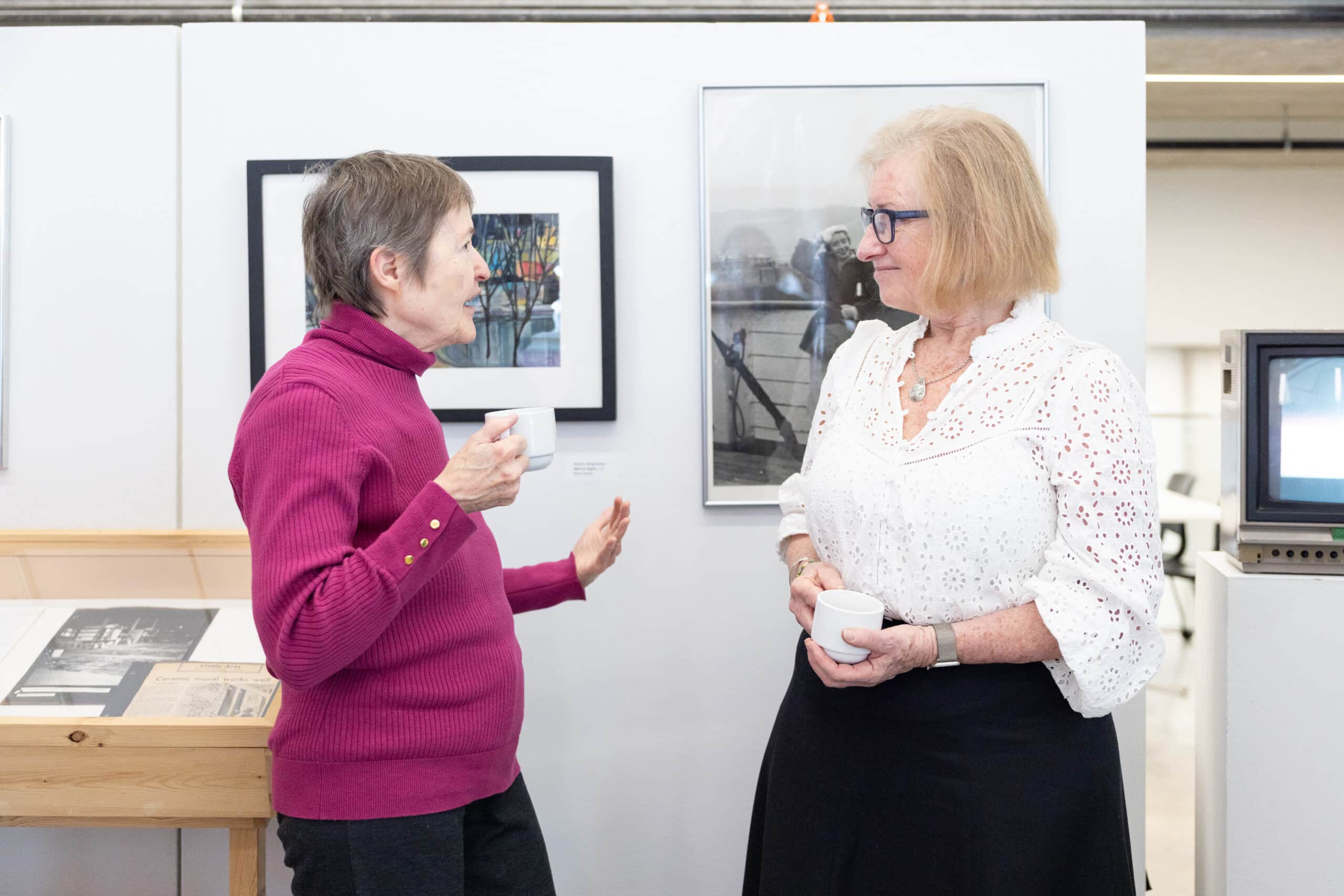 Two visitors talk face to face while holding cups in a gallery, with framed photographs displayed on the wall behind them.