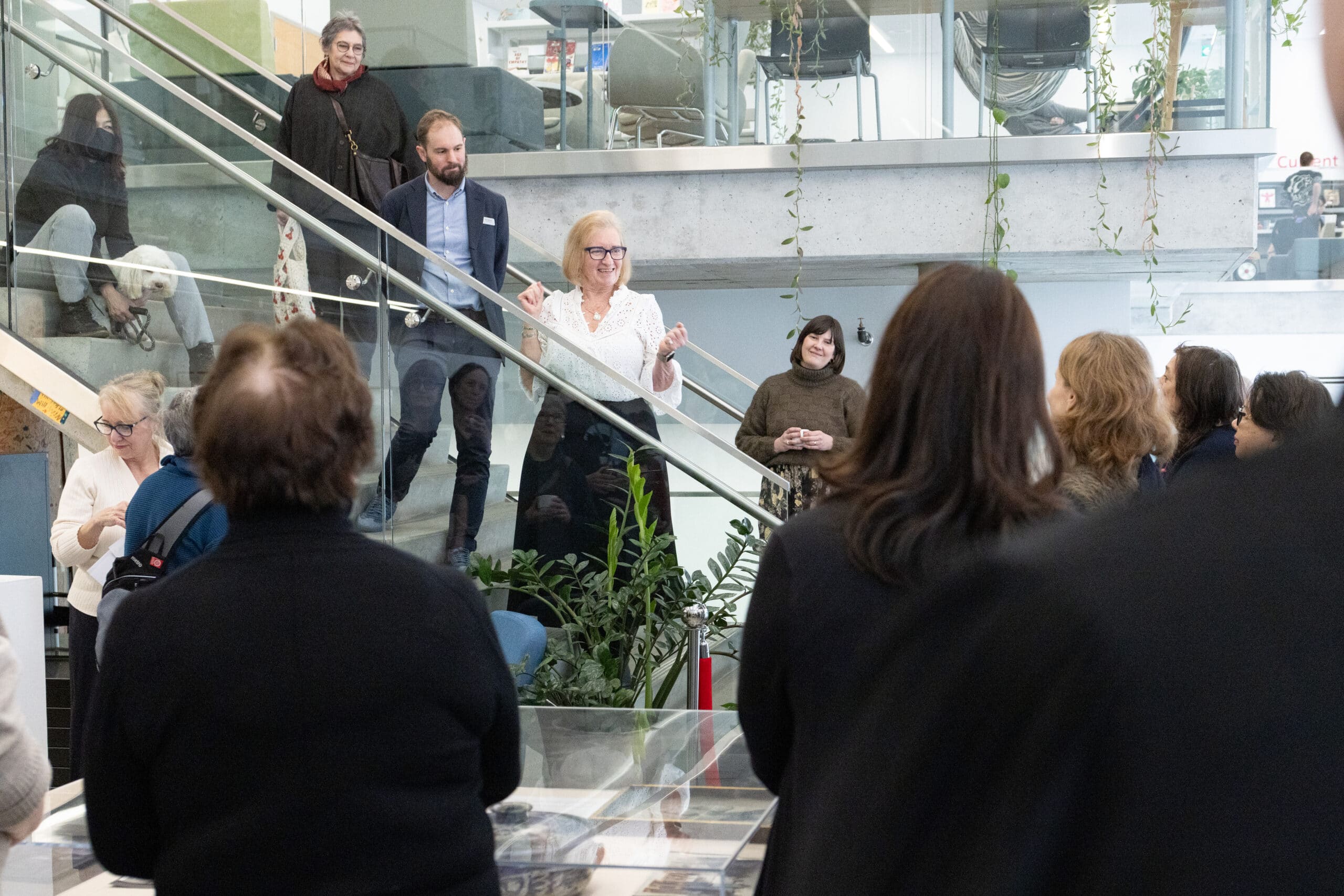 A person stands on a staircase addressing a group of visitors gathered below in a gallery during an exhibition talk.