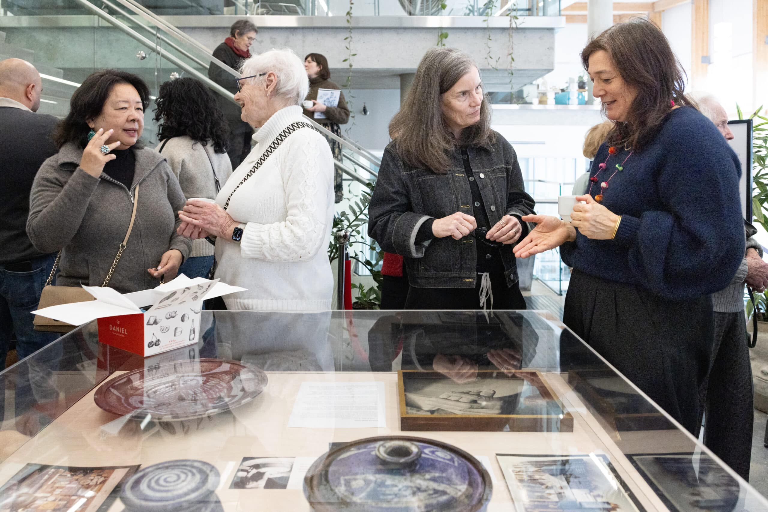 Small groups of visitors converse beside a glass display case filled with ceramics and images in a bright exhibition space.