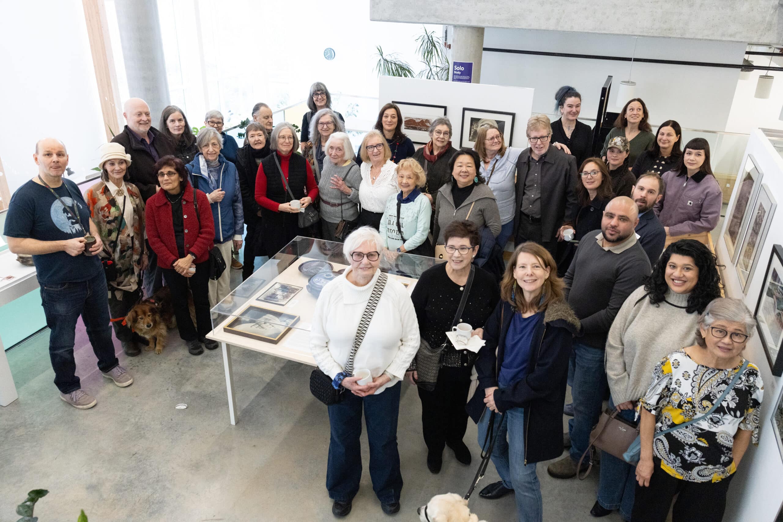 A large group of visitors gathers around a display case in a gallery, posing together among framed artworks and exhibition displays.