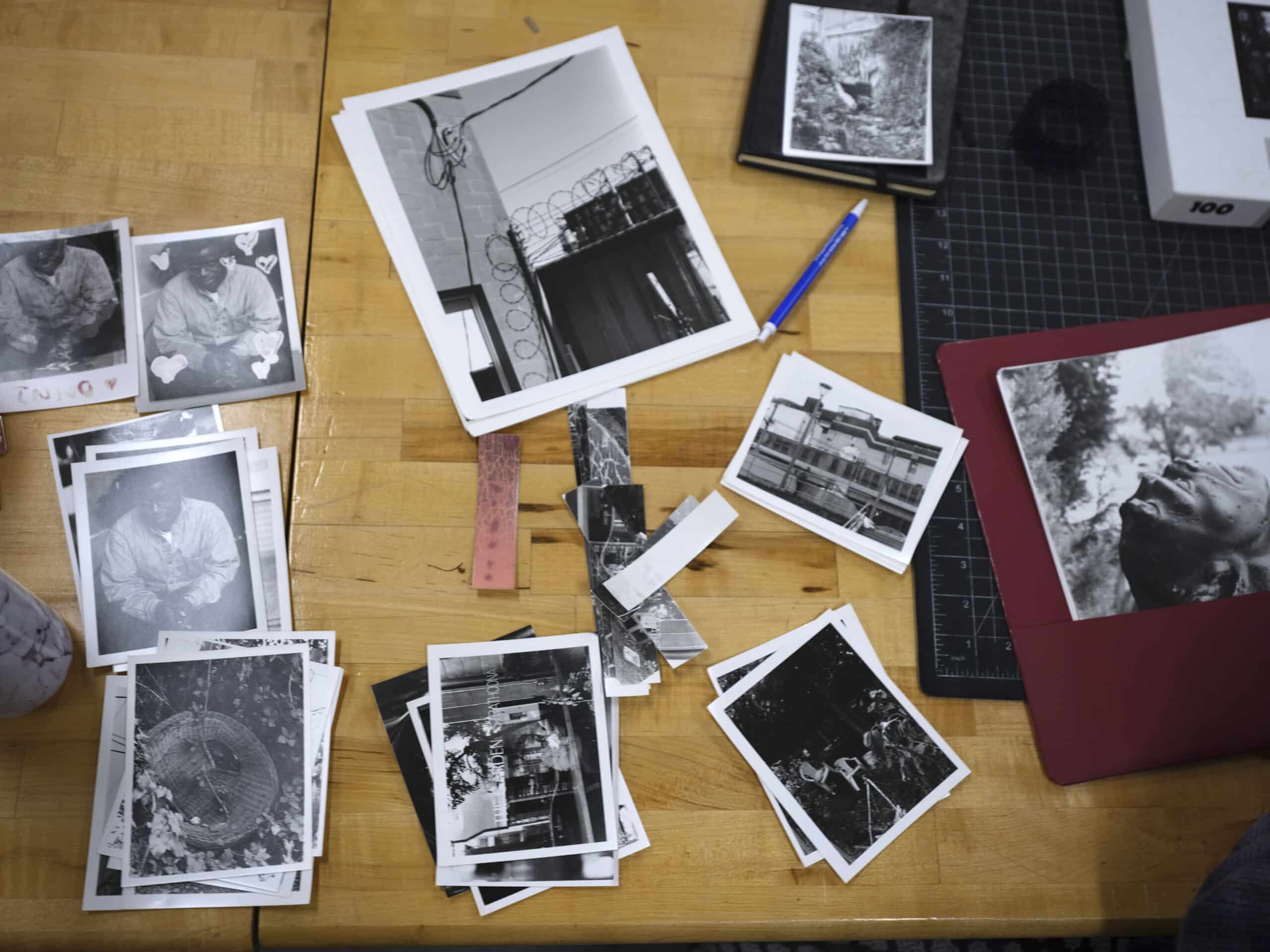 Overhead view of a wooden worktable covered with scattered black-and-white photo prints. Small stacks of photographs show portraits, buildings, and outdoor scenes, alongside strips of film negatives. A blue pen, a black cutting mat, a closed notebook, and a red portfolio holding a large portrait print sit among the images, suggesting an active photo editing or selection process.