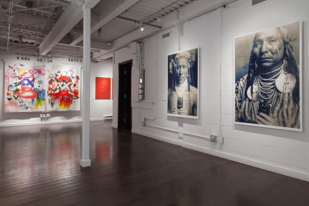 A spacious gallery opens onto multiple works: vivid abstract paintings on one wall and large portrait photographs on another, their faces and forms holding the room in a calm, reflective hush.