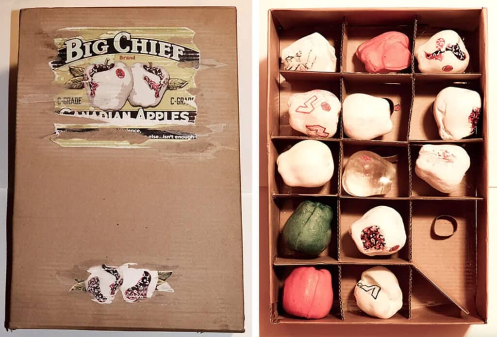 A worn cardboard fruit box labeled “Big Chief apples” sits beside an open tray of small, hand-painted objects arranged in neat compartments, each piece carrying marks, colors, and quiet variation.