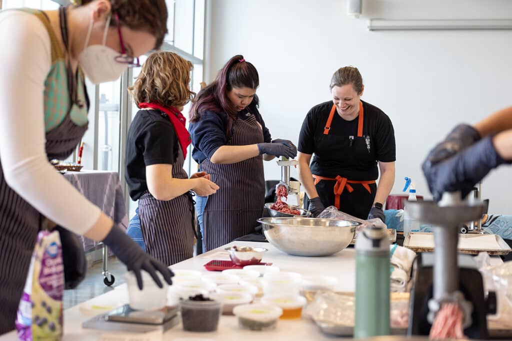 Several people gather around a bright worktable, hands busy measuring, mixing, and guiding meat through a grinder. Small containers, bowls, and tools scatter the surface, holding a shared rhythm of preparation and care.