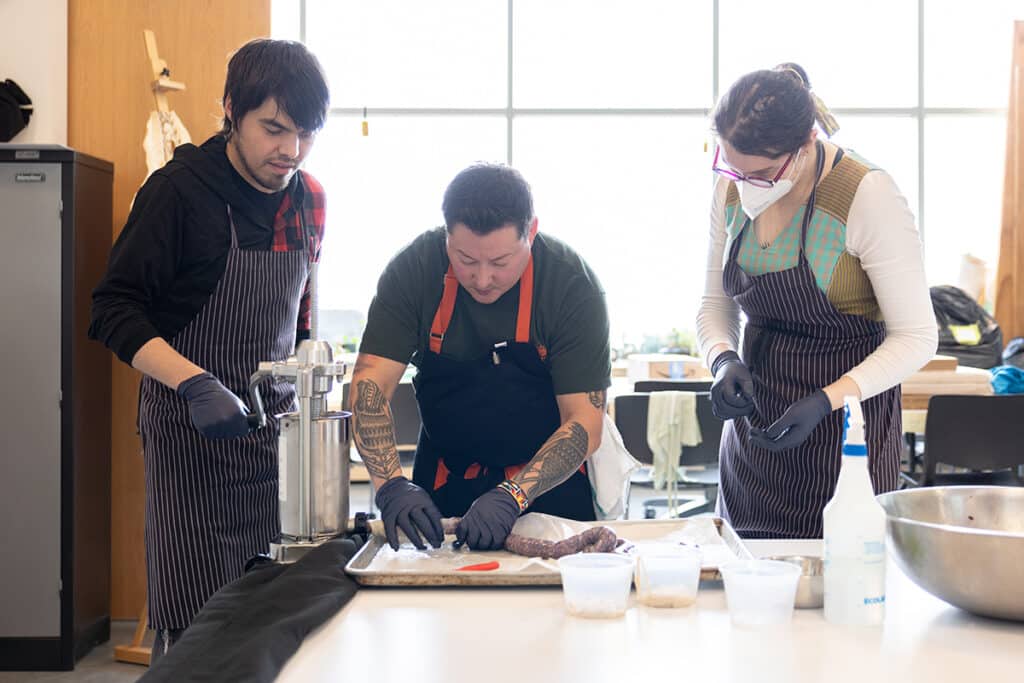 Three people stand close at a table, shaping and filling sausage together. Their gloved hands move with focus, framed by soft window light and a quiet sense of collaboration.
