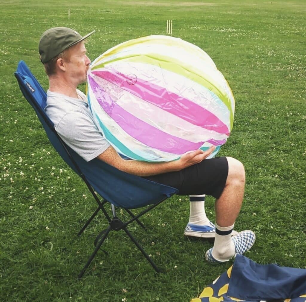A person reclines in a folding chair on green grass, holding a striped inflatable ball close, a moment of quiet play and pause in an open field.