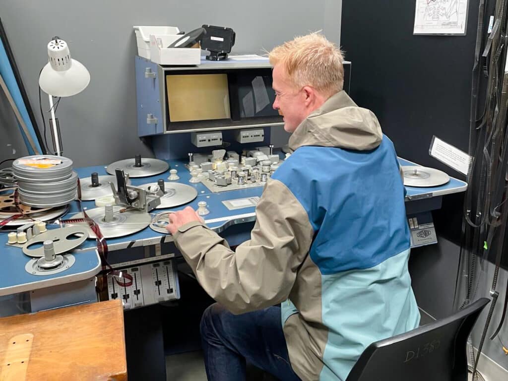 A person sits at a film editing bench, hands guiding reels and dials, surrounded by spools, light, and the quiet mechanics of moving images.