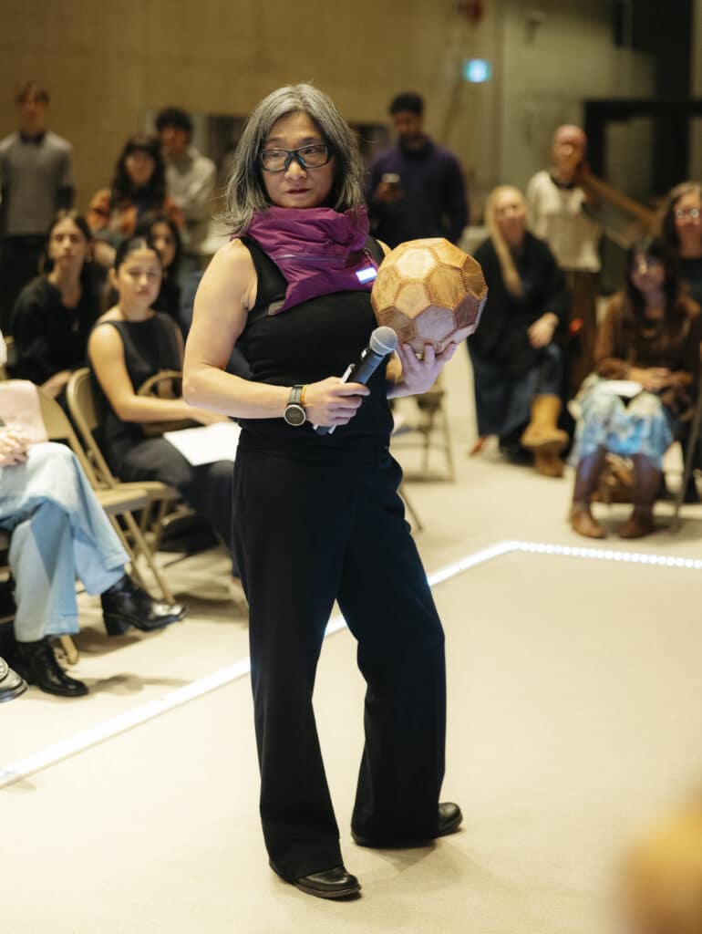 A person stands on a runway before an audience in dark clothing with a magenta scarf and sunglasses holding a geometric orb and a microphone. 