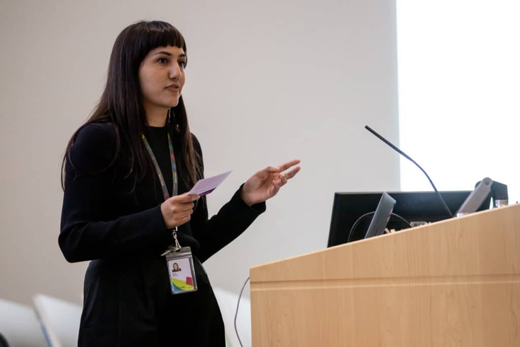 A person stands at a podium, holding a small card and gesturing mid-speech; a microphone and screen sit nearby as their focused expression and steady posture carry their words into a quiet, attentive room.