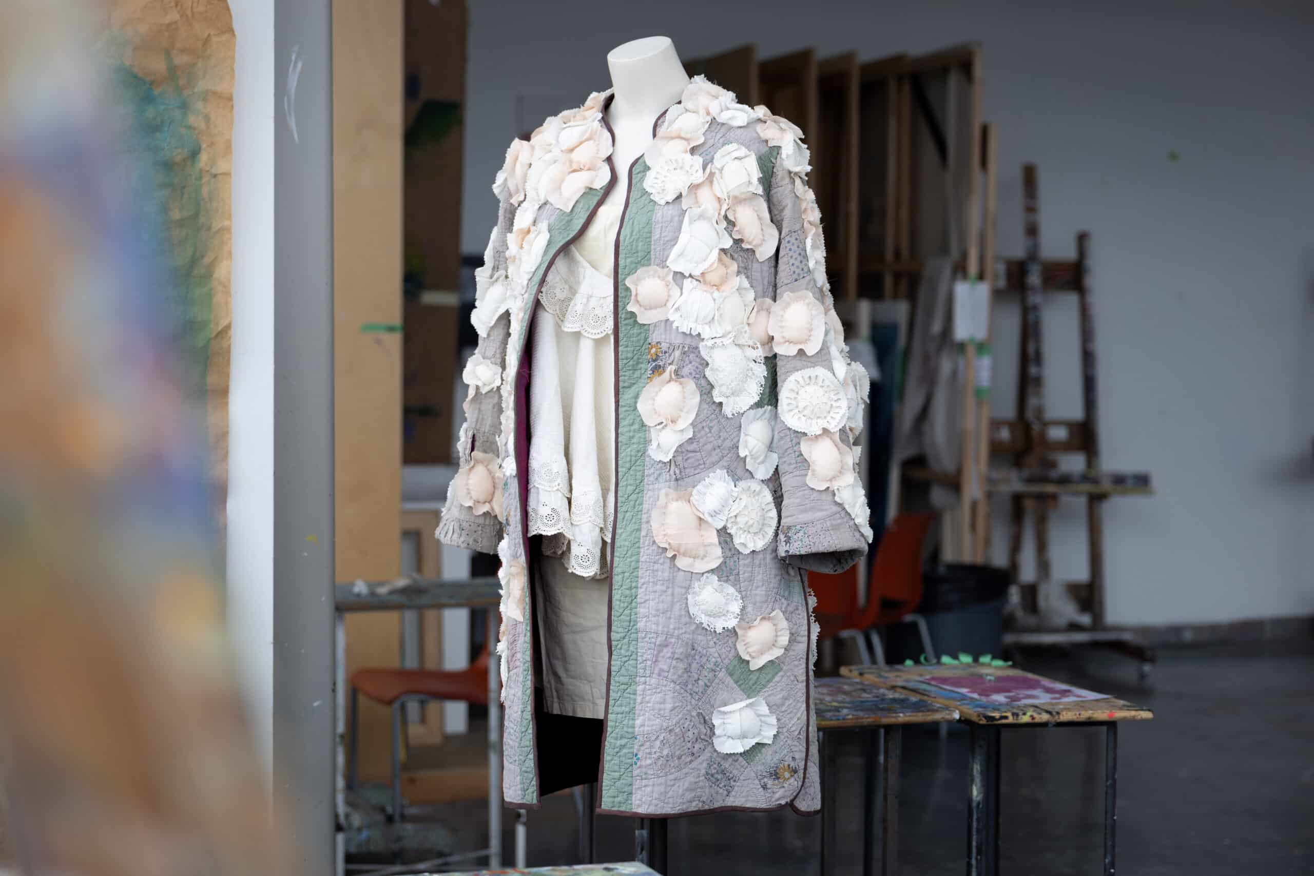 A handmade coat with textile blossoms resembling flowers is illuminated by soft light from a window.