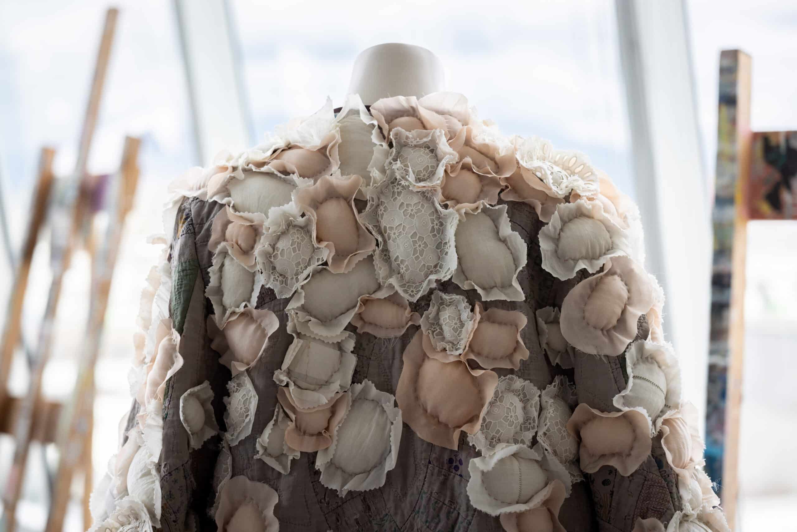 A handmade coat with textile blossoms resembling flowers is illuminated by soft light from a window.