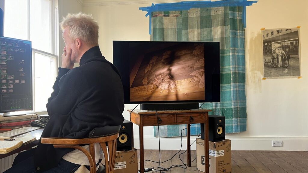 A person sits at a desk by a window, editing on a computer while a screen nearby shows a softly lit scene; cables, speakers, and taped fabric frame a space of focus and making.
