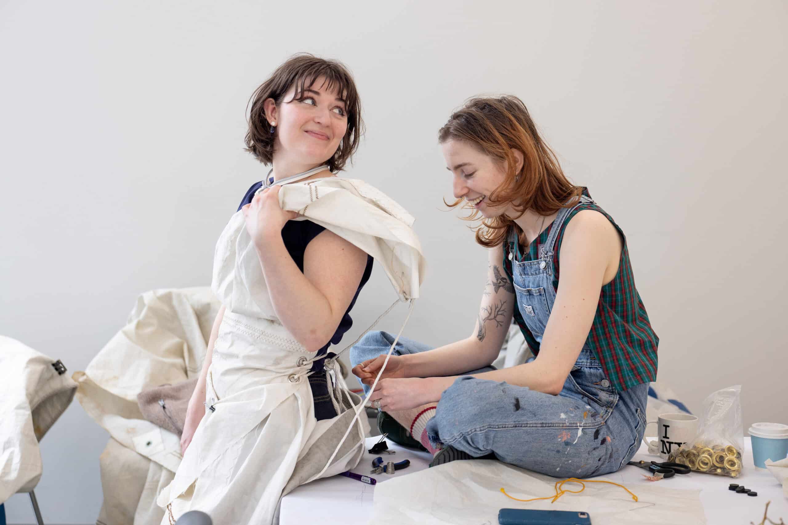 Two people sit close together on a worktable, smiling as one adjusts a softly draped white garment, the fabric gathered like a quiet collaboration.