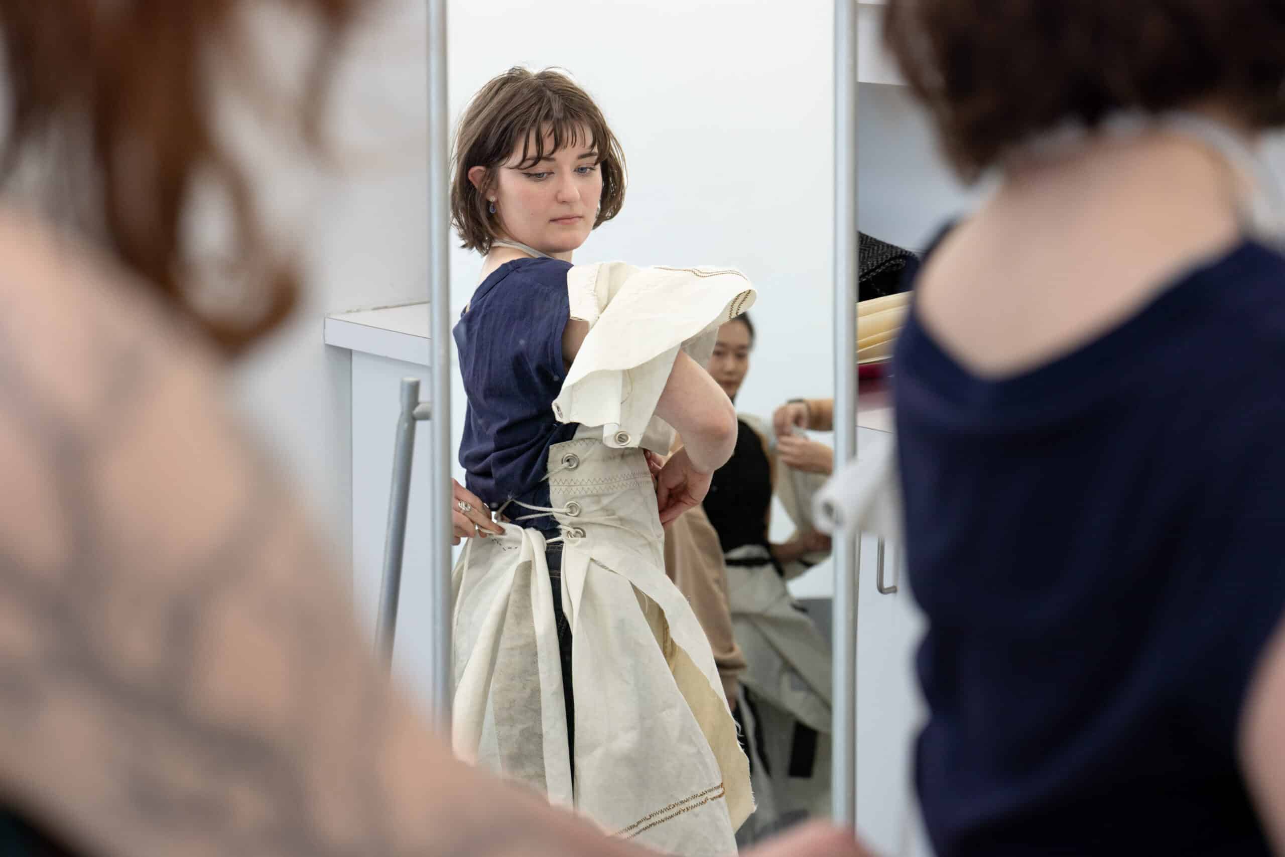 A person studies their reflection while fastening a structured, laced garment, the mirror holding both focus and quiet concentration.