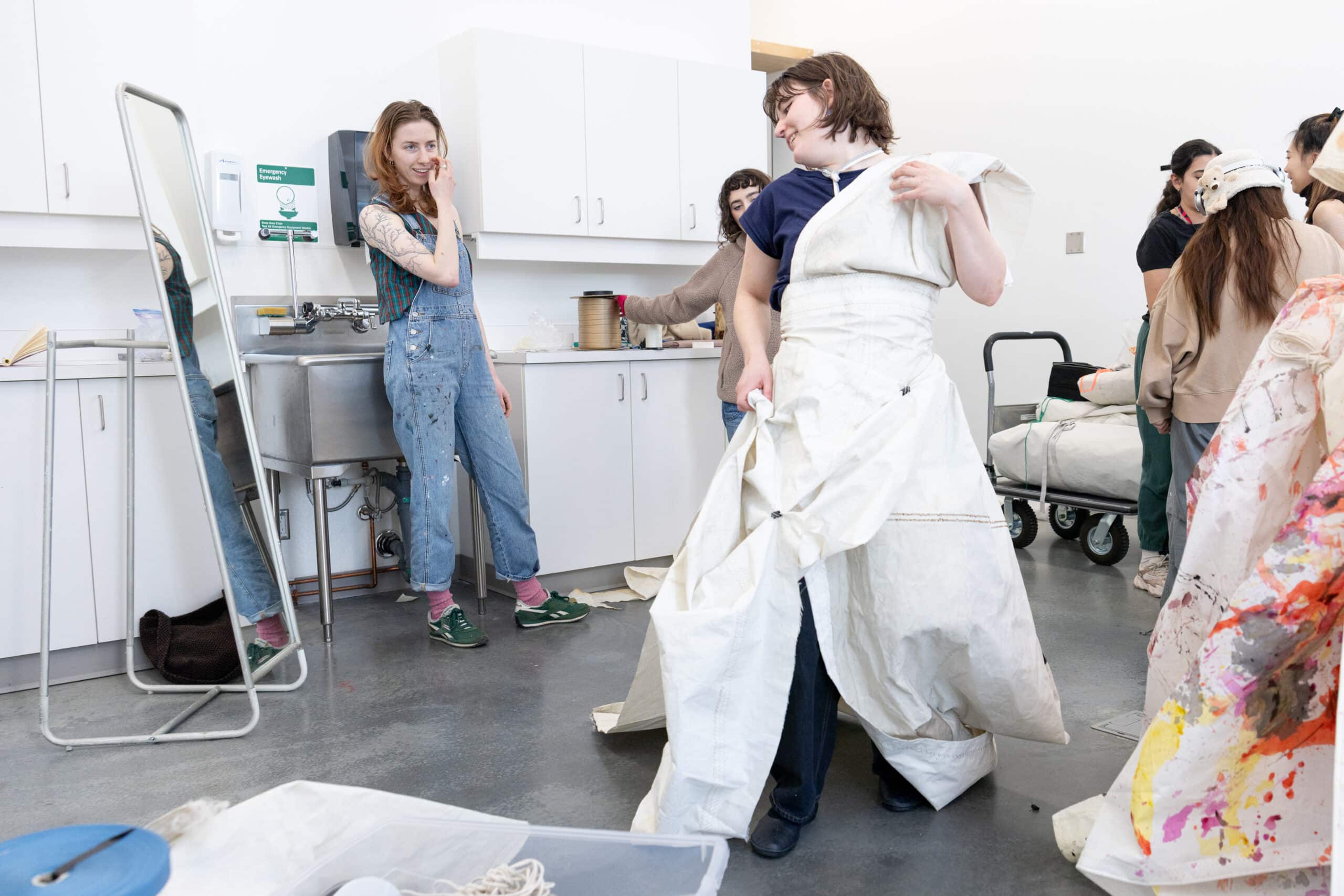 In a bright studio, a person turns in a voluminous white piece as others watch and prepare materials, the room alive with making.